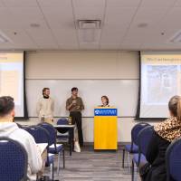 three students presenting at the front of a carpeted conference room